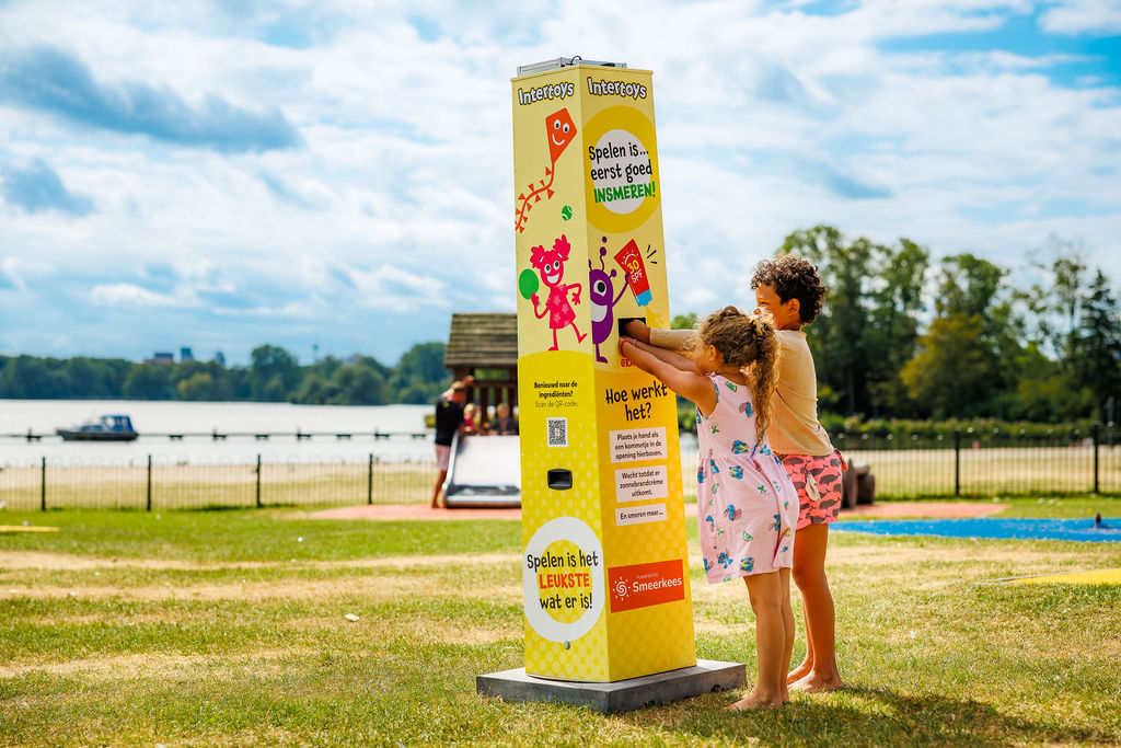 Twee kinderen halen zonnebrandcrème bij een zonnebrandcrème-station in Intertoys-stijl op een speelplaats aan het meer