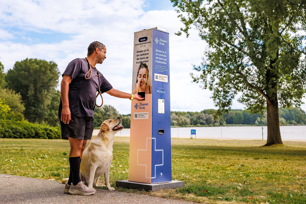 Man met hond bij een zonnebrandcrème-stand van de Nederlandse zorgverzekeraar Zilveren Kruis in een park aan het meer