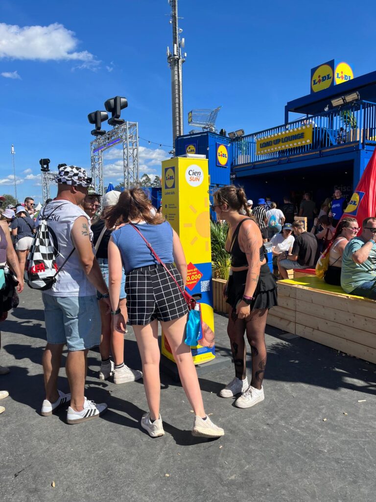 Jonge festivalgangers staan bij een gele zonnebrandcrème-dispenserkolom van Lidl en Cien Sun voor de Lidl „Load Up Lounge” op een zonnig festivalterrein